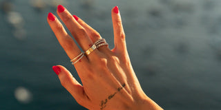 Hand raised against the ocean wearing stacked silver and gold IKAIKA rings, red nails