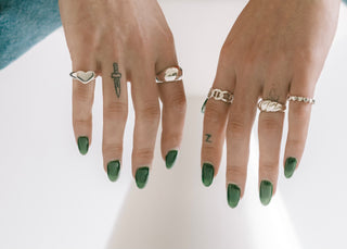 Two hands with green nails wearing a heart signet, chain rings and stacking rings